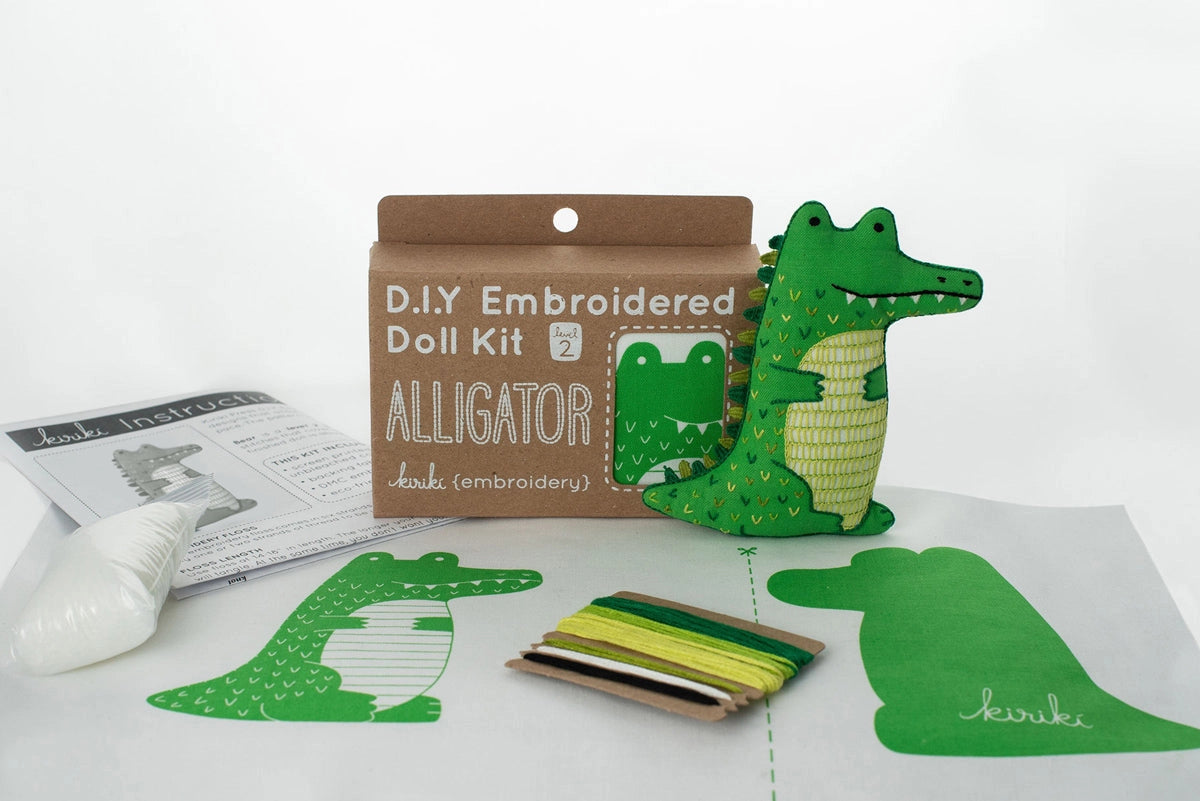 The Faire Alligator Embroidery Kit includes green and white thread, pre-cut fabric with an alligator pattern, stuffing, instructions, and a finished sample—everything needed to craft your own alligator plush.