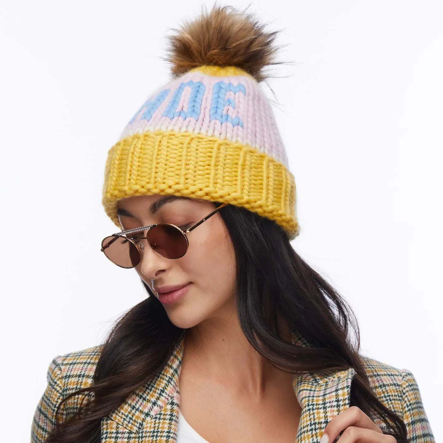A woman in round sunglasses and a Faire Knit Ski Hat with a brown faux fur pom pom, paired with a plaid jacket, looks down slightly with a neutral expression against a white background.