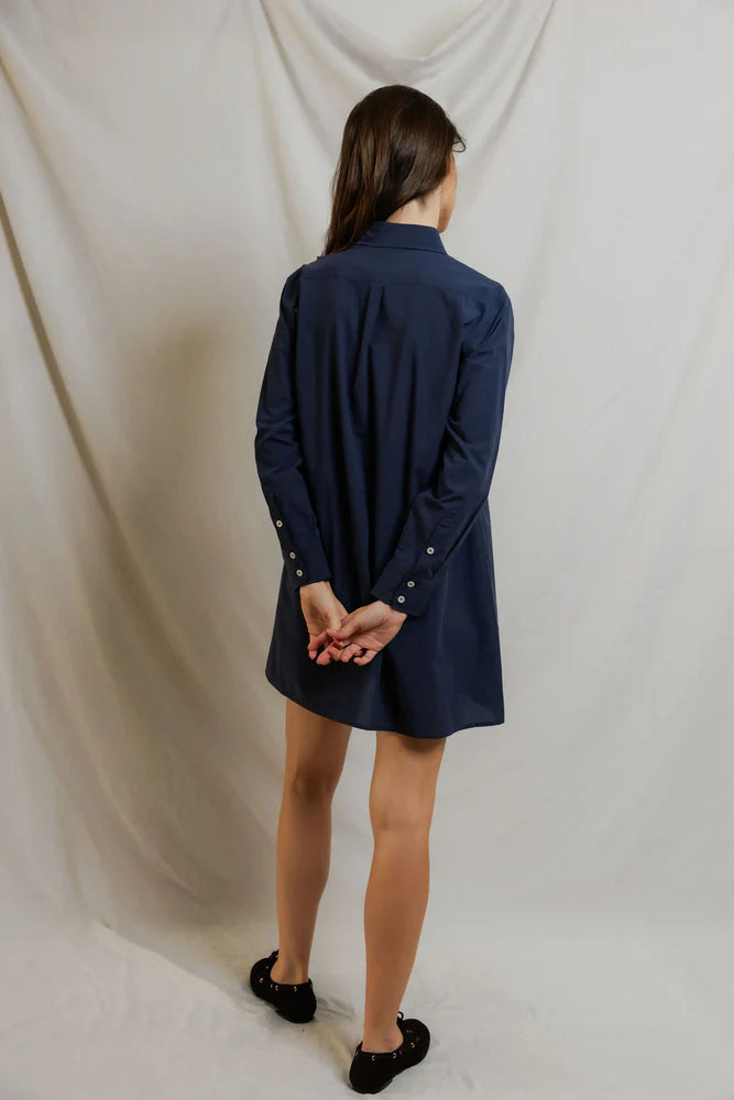 A woman faces away, wearing the Felicia Poplin Dress by Perfectwhitetee in navy with black flats. Her arms are behind her back as she poses against a plain light backdrop—an effortless pick for everyday wear.