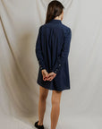 A woman faces away, wearing the Felicia Poplin Dress by Perfectwhitetee in navy with black flats. Her arms are behind her back as she poses against a plain light backdrop—an effortless pick for everyday wear.
