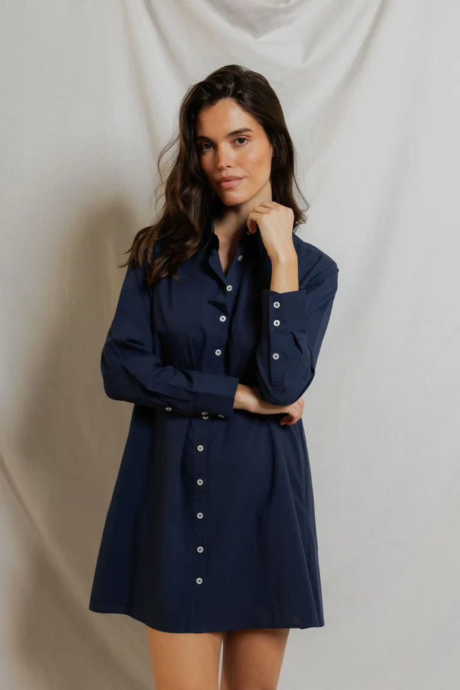 A woman with long dark hair models the Felicia Poplin Dress by Perfectwhitetee in navy blue, standing before a light backdrop with her chin resting on her hand—an effortless everyday look.
