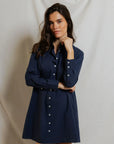 A woman with long dark hair models the Felicia Poplin Dress by Perfectwhitetee in navy blue, standing before a light backdrop with her chin resting on her hand—an effortless everyday look.