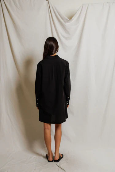 A woman with long hair stands facing away, wearing the Perfectwhitetee Felicia Poplin Dress and black sandals against draped white fabric—an ideal choice for effortless everyday style.