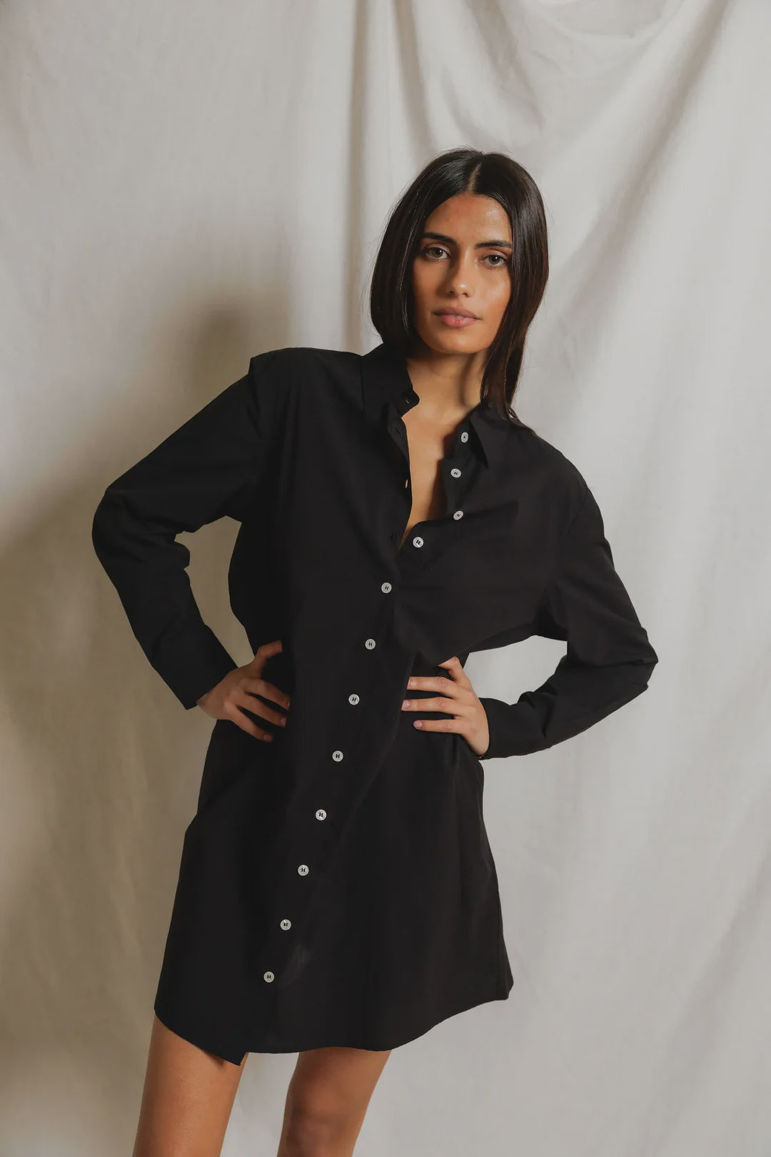 A woman poses in front of a light backdrop, wearing the long-sleeved Felicia Poplin Dress by Perfectwhitetee—a black cotton poplin shirt dress with white buttons—ideal for everyday wear.