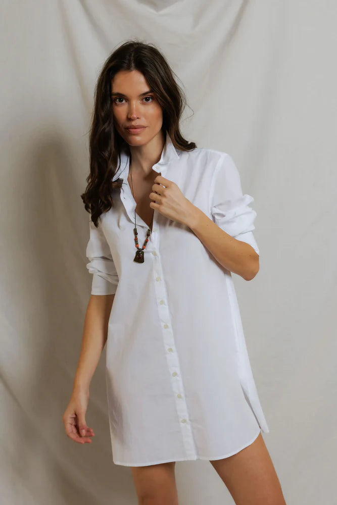 A woman with long brown hair stands before a light backdrop, wearing the Felicia Poplin Dress by Perfectwhitetee with rolled sleeves and a brown pendant necklace—ideal for effortless everyday style.