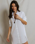 A woman with long brown hair stands before a light backdrop, wearing the Felicia Poplin Dress by Perfectwhitetee with rolled sleeves and a brown pendant necklace—ideal for effortless everyday style.