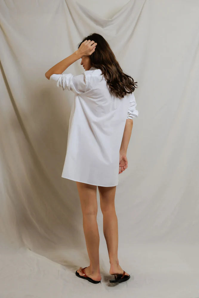 A woman with long, wavy brown hair faces away in the Felicia Poplin Dress by Perfectwhitetee—an oversized white cotton shirt dress paired with black flip-flops, set against a neutral backdrop. Ideal for effortless, everyday style.