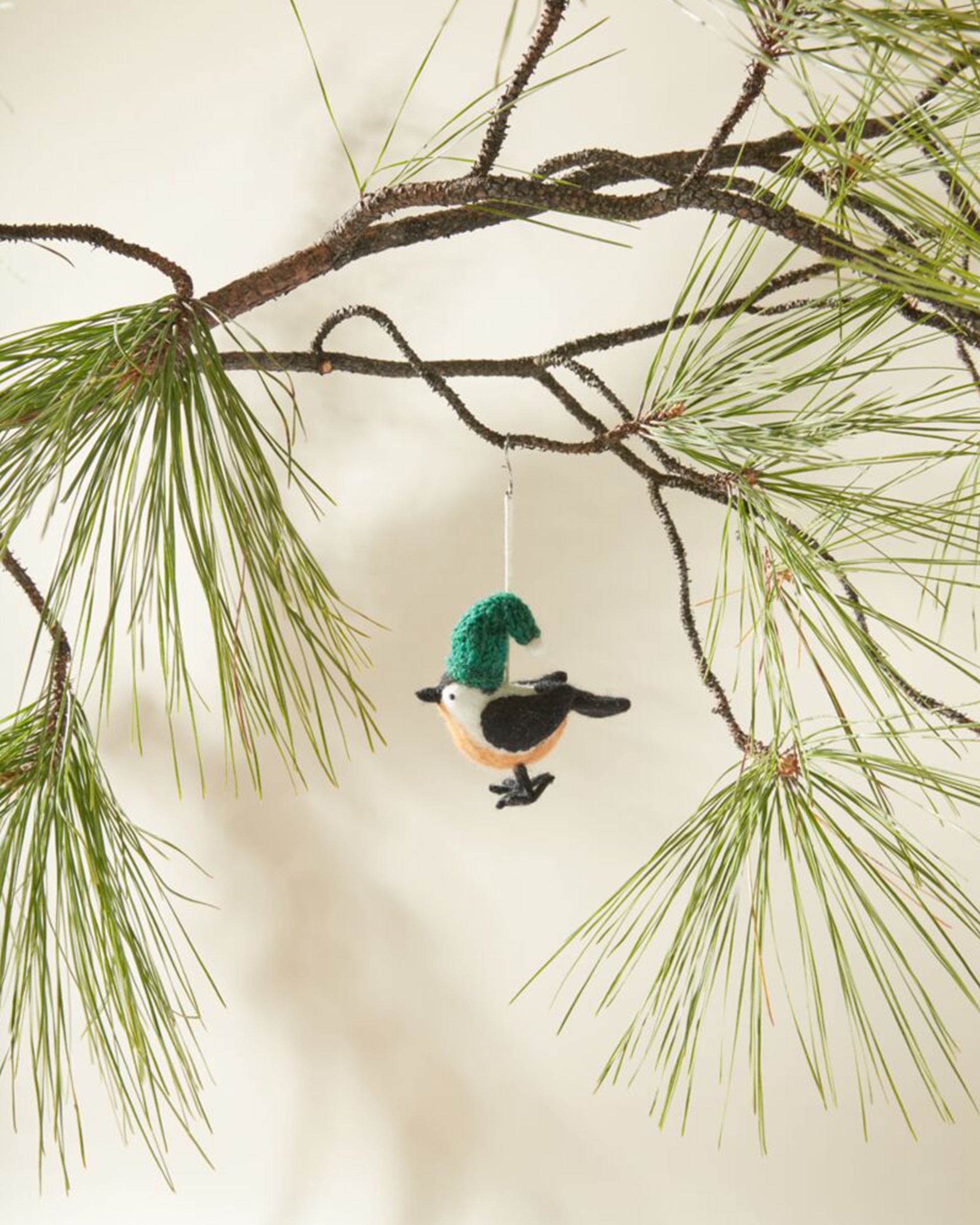 The Accent Decor Cozy Weather Ornament, a small felt bird with a green hat, hangs from a pine branch with long needles against a plain, light background.