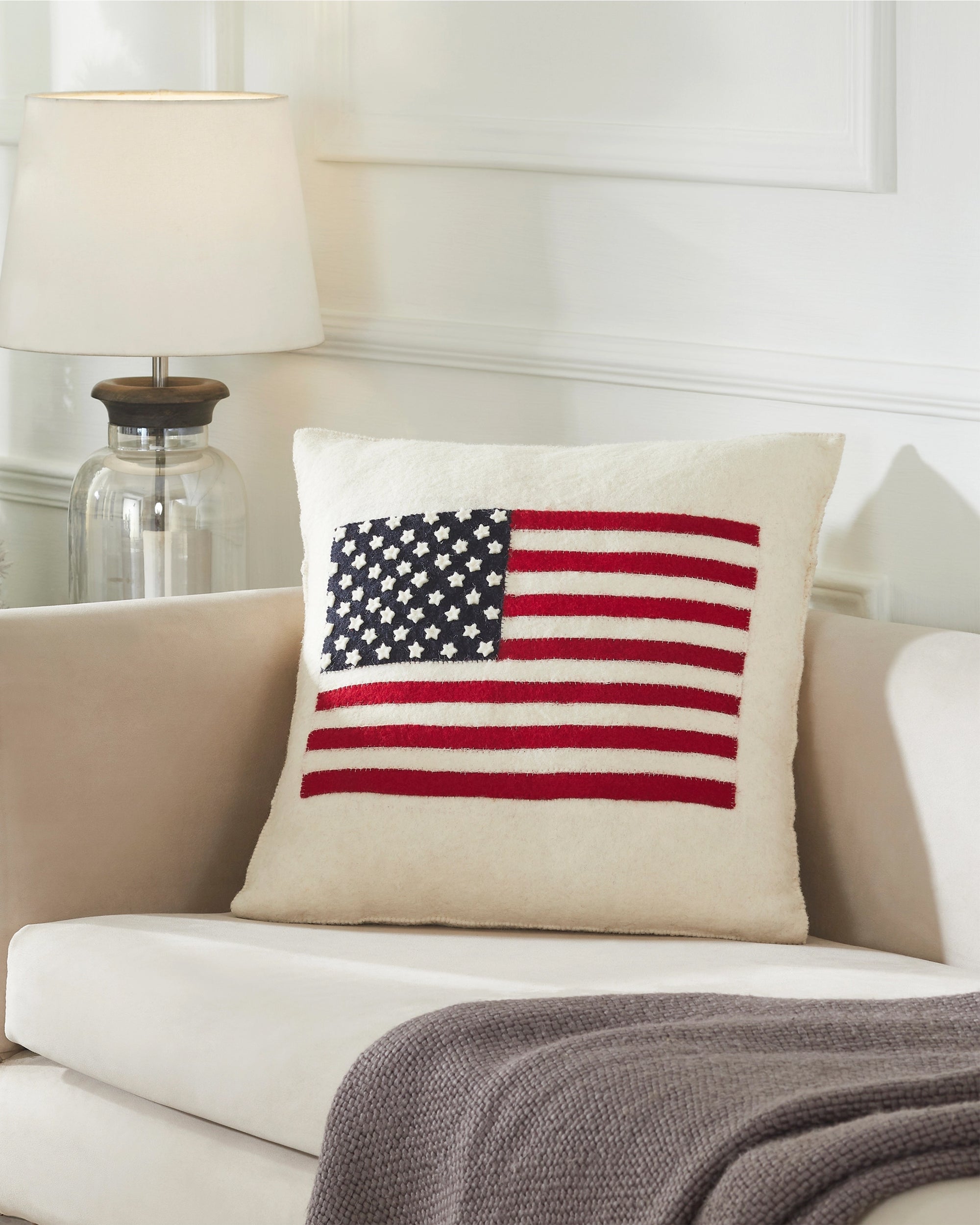 The Faire American Flag Wool Pillow, in cream felted wool and hand-embroidered, sits on a beige sofa beside a gray blanket, with a glass-based lamp visible on a table in the background.