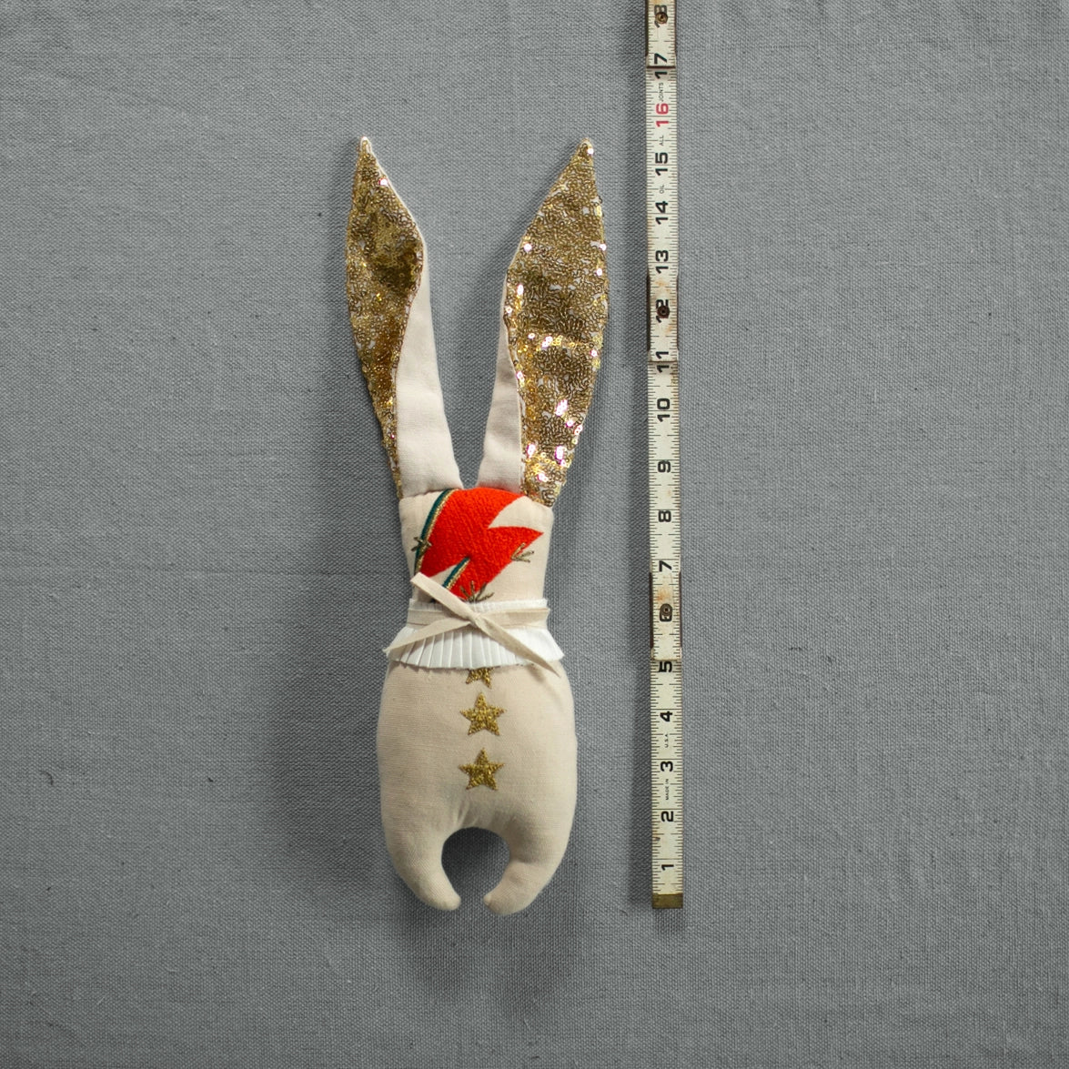 The Bowie Bunny by Faire is a fabric rabbit toy inspired by David Bowie, featuring long ears with a gold pattern and holding a red carrot. It sits next to a measuring tape on a grey background to display its size and is adorned with star decorations, making it an enchanting art doll for whimsical decor.