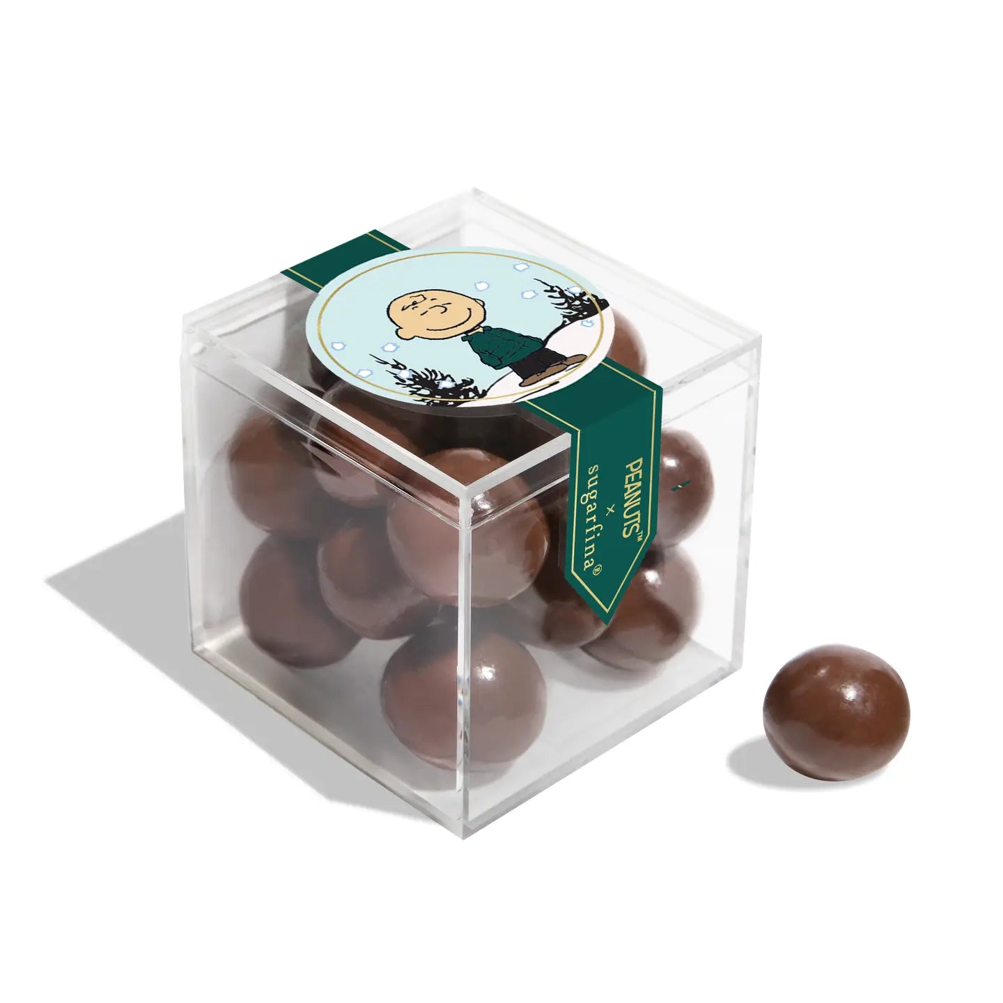 The Faire Christmas Candy Cube is a clear box of chocolate-covered balls with a Charlie Brown Peanuts sticker on the lid—one ball outside. Ideal for sharing assorted candies or gifting as cheerful holiday sweets.