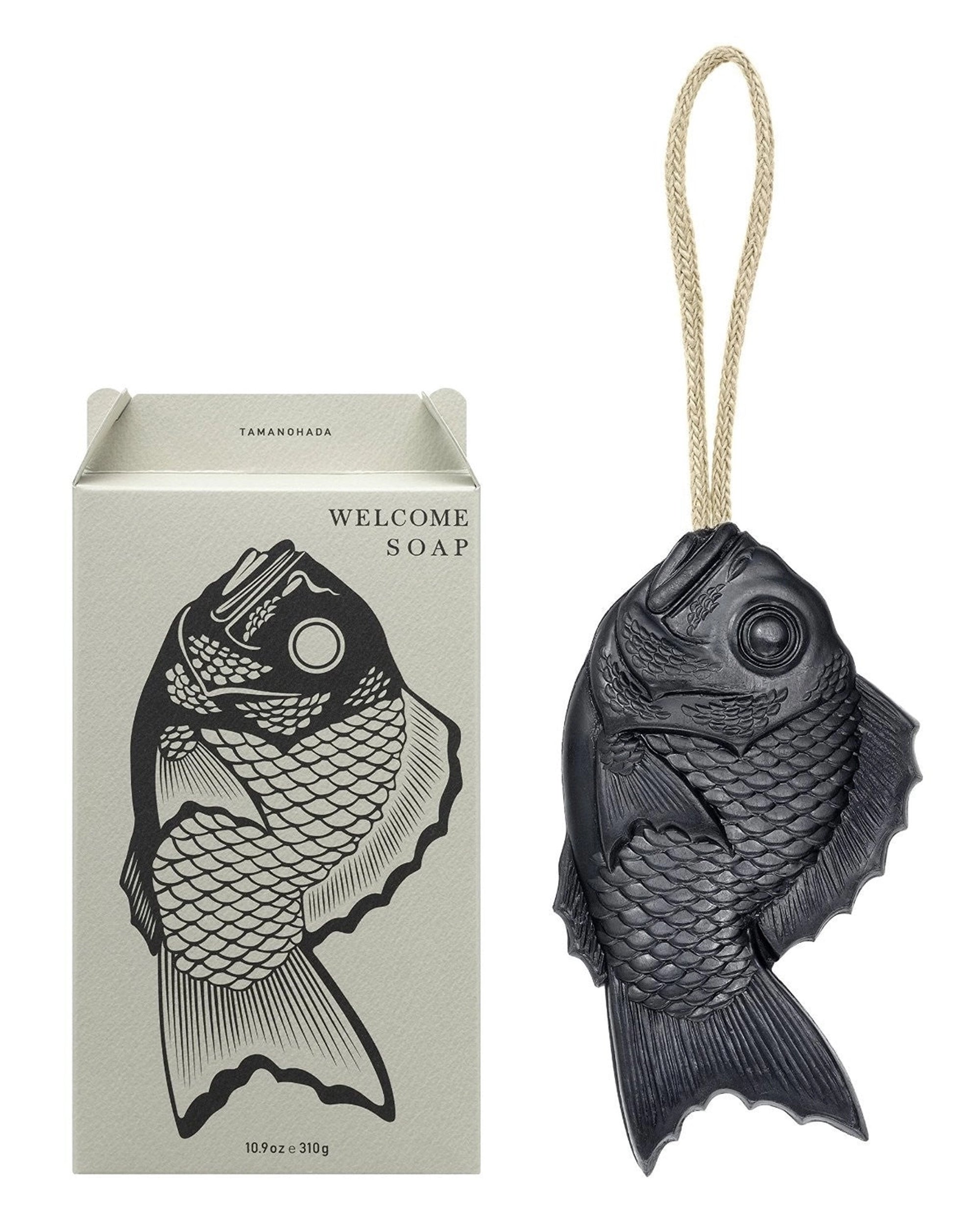 The Faire Tamanohada Soap, shaped like a black fish and featuring a hanging rope, is shown beside its box with a matching fish illustration and "WELCOME SOAP" text. This elegant soap is crafted from plant-based ingredients.