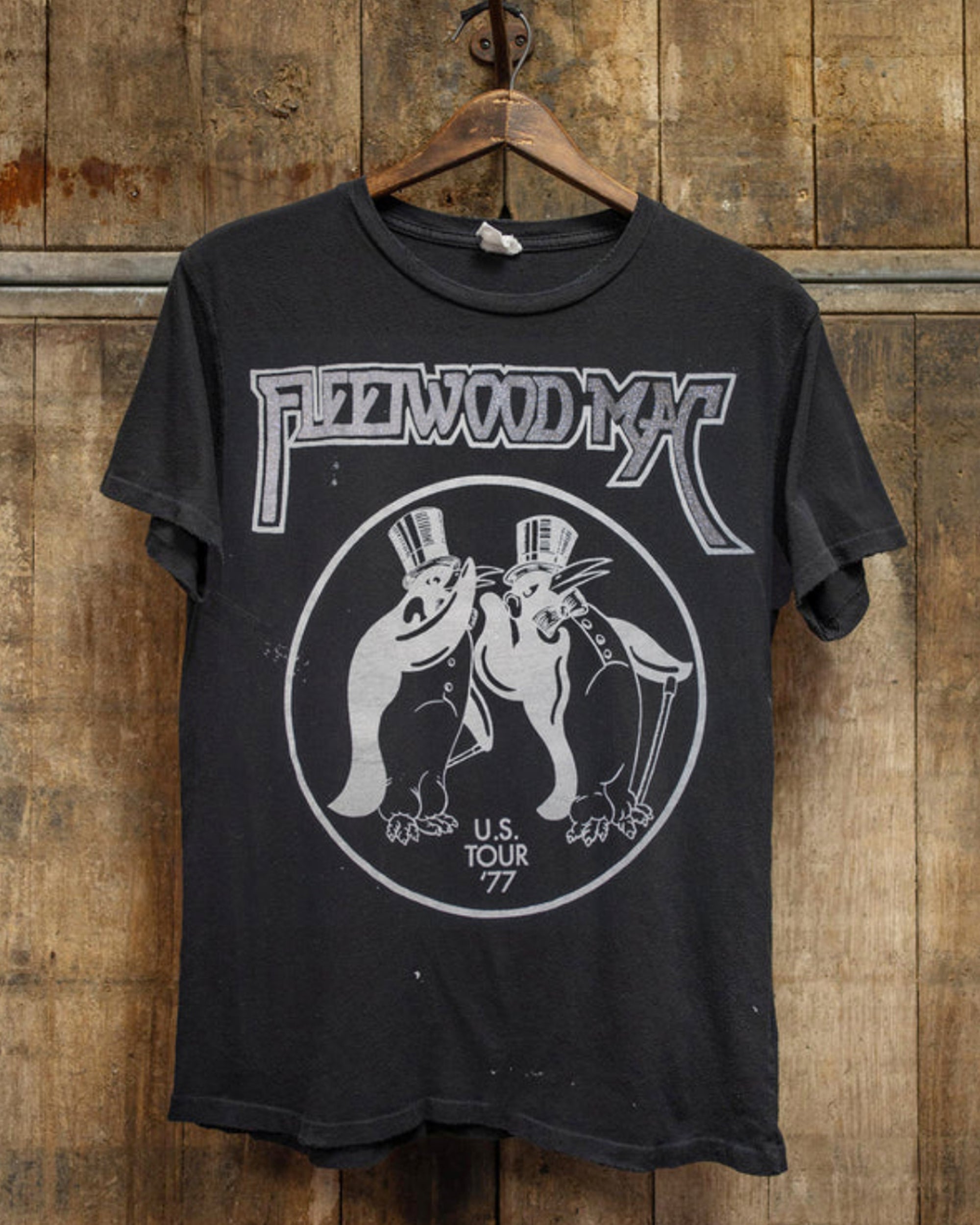 The Made Worn Fleetwood Mac Tee is a vintage unisex black t-shirt with two penguins in top hats and "U.S. Tour '77" under the band's logo, made in the USA and displayed on a wooden hanger against a rustic wood wall.