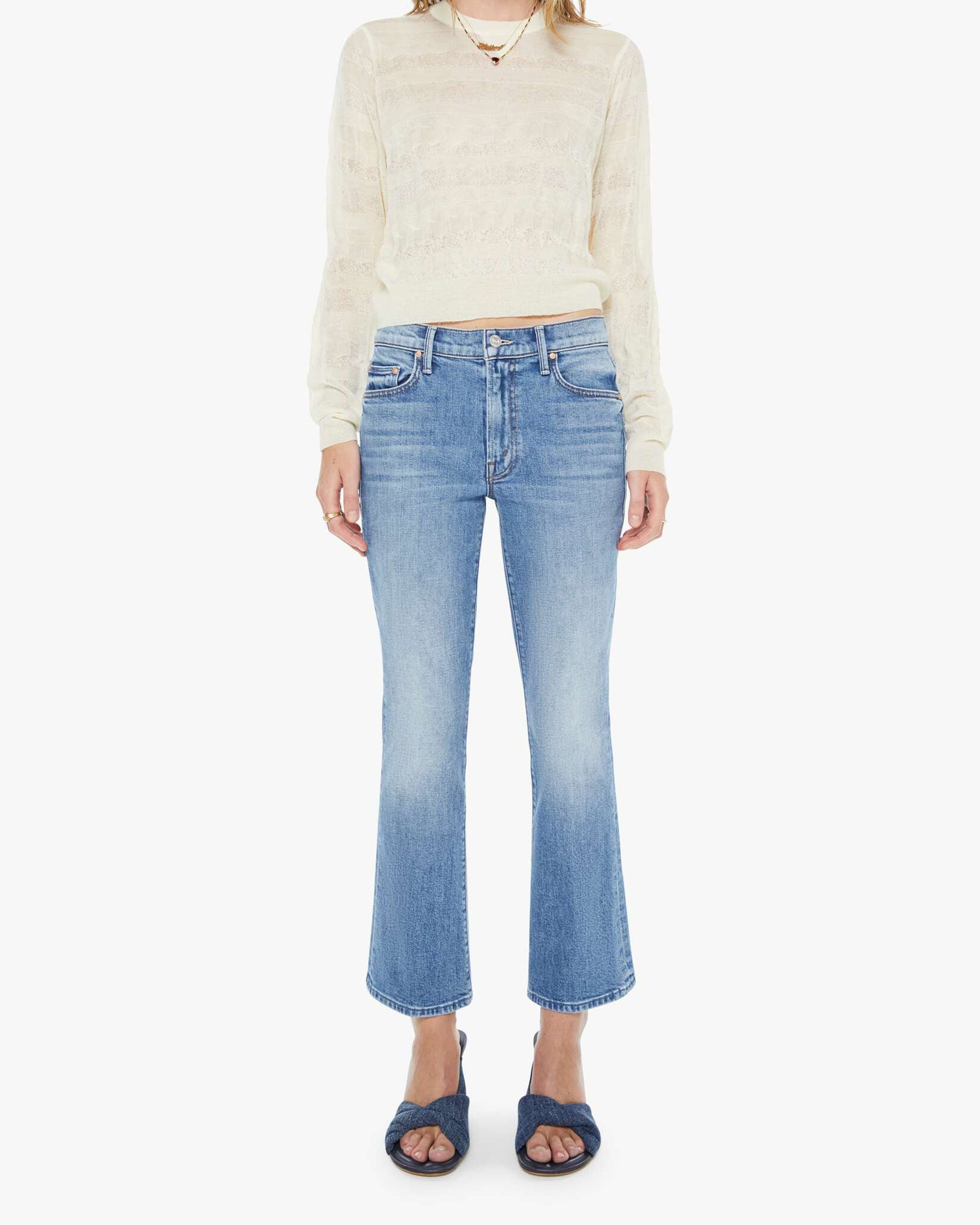 A person stands against a white background wearing The Outside Flood cropped flare vintage blue jeans by Mother, a sheer cream long-sleeve sweater, and dark blue open-toe sandals.