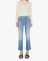 A person stands against a white background wearing The Outside Flood cropped flare vintage blue jeans by Mother, a sheer cream long-sleeve sweater, and dark blue open-toe sandals.