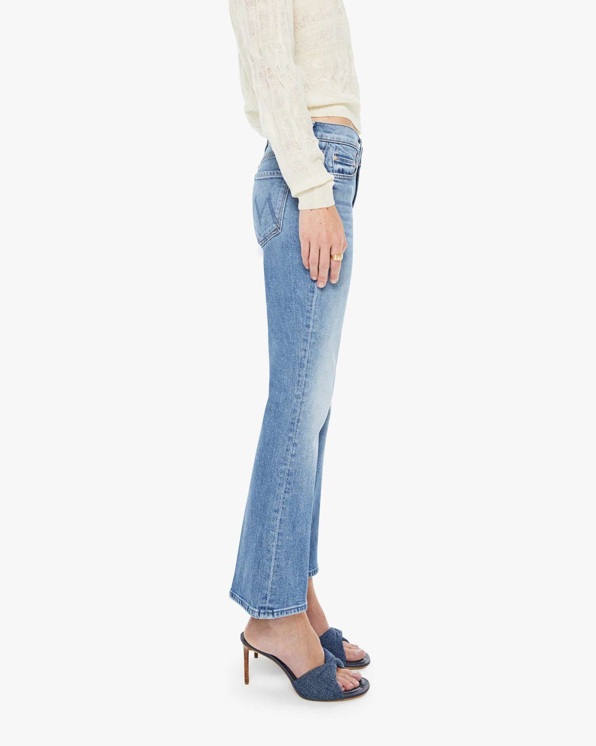A person stands sideways against a white background, wearing Mother’s The Outside Flood vintage blue cropped flare jeans, a cream long-sleeve top, and blue heeled sandals.
