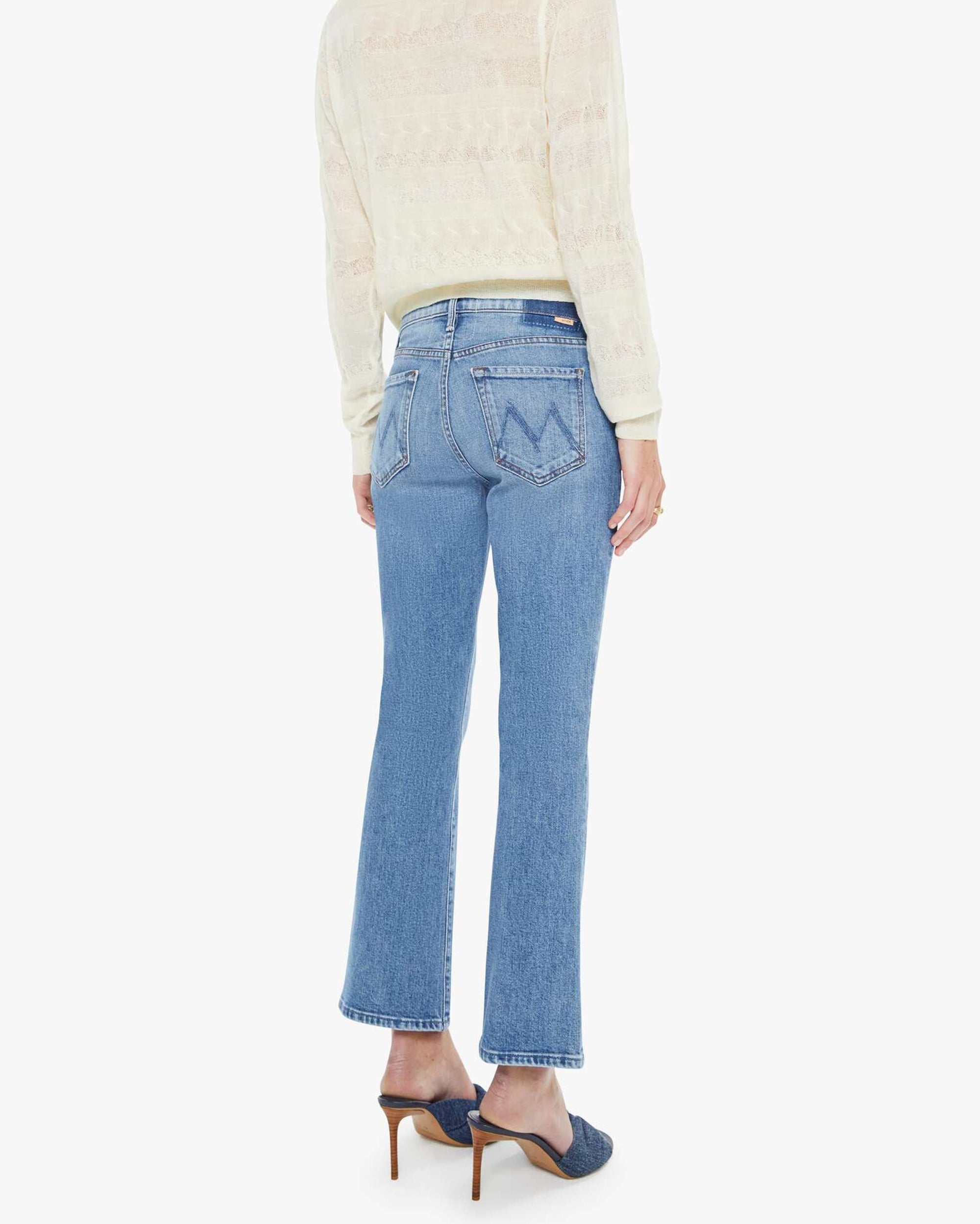 A person wearing Mother’s The Outside Flood vintage blue straight-leg jeans and a sheer cream sweater stands facing away, also wearing blue heeled mules.