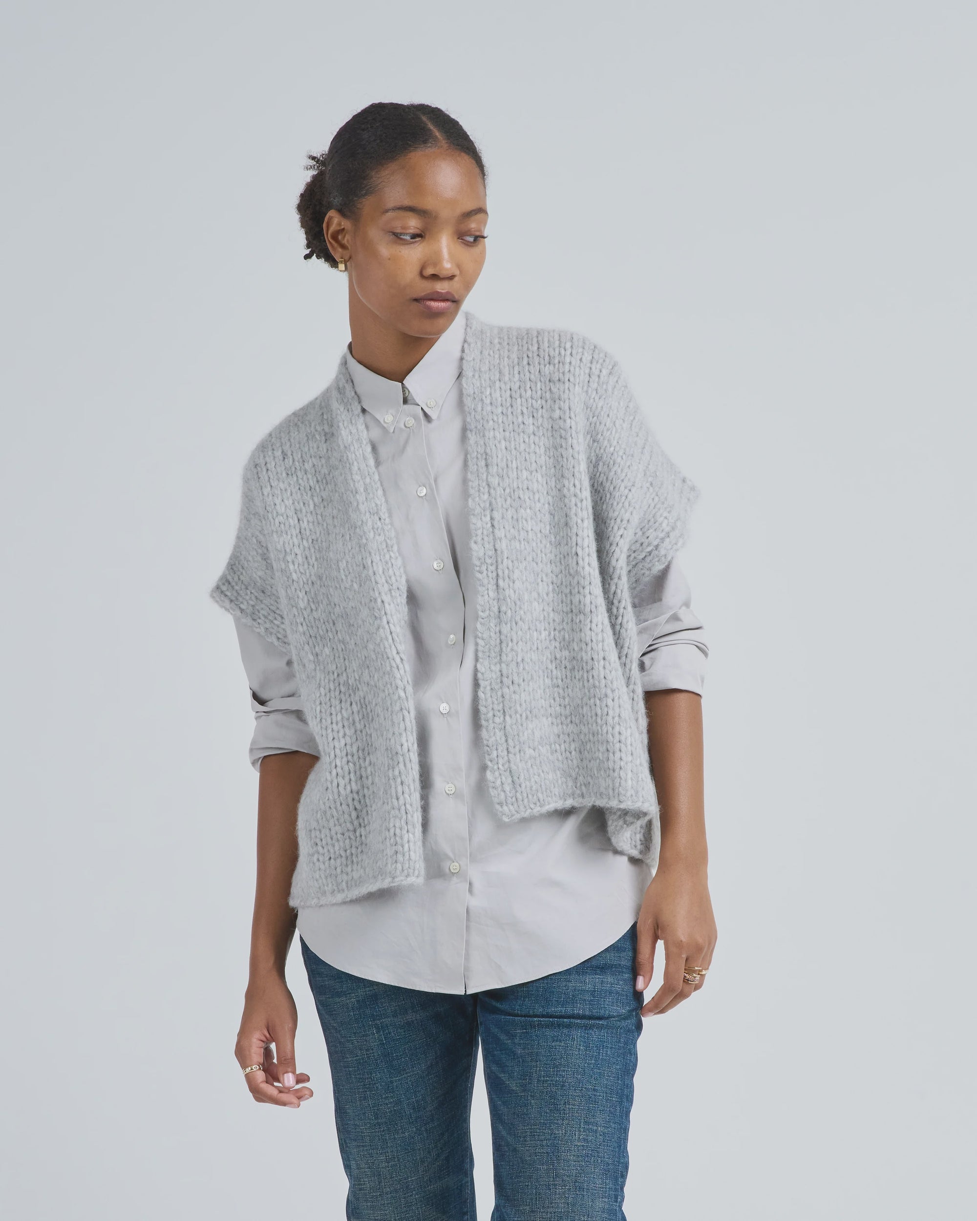 Wearing the LaTierra Flor Vest—a loose, chunky-knit, light gray merino wool vest—over a light gray button-up and blue jeans, a person stands against a plain background, gazing downward with a neutral expression.