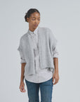 Wearing the LaTierra Flor Vest—a loose, chunky-knit, light gray merino wool vest—over a light gray button-up and blue jeans, a person stands against a plain background, gazing downward with a neutral expression.