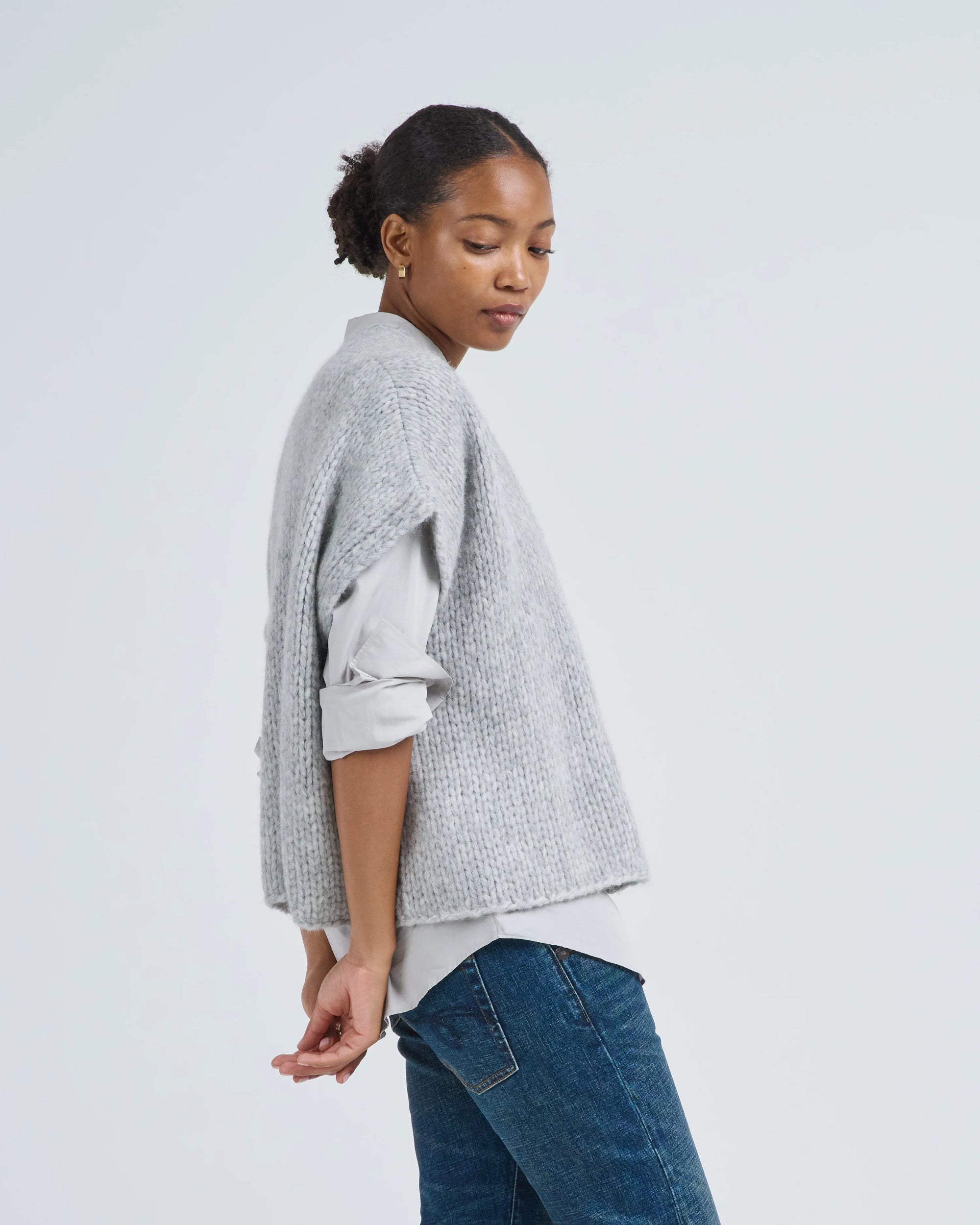 A woman stands sideways, looking down, wearing the Flor Vest by LaTierra—a loose light grey merino wool knit—layered over a long-sleeve shirt and blue jeans against a plain light background.