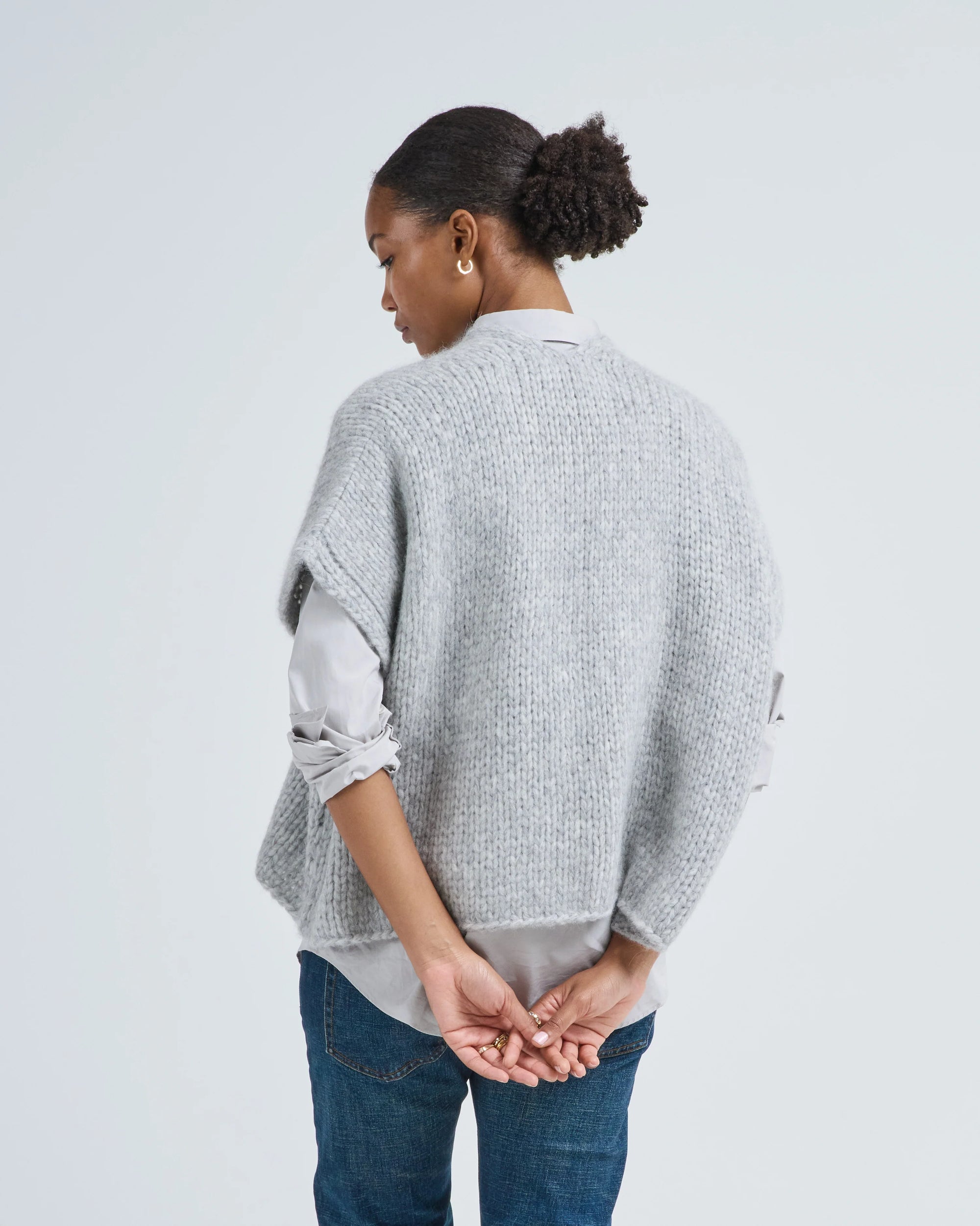 A woman with her hair in a bun stands with her back to the camera, wearing LaTierra’s Flor Vest—a light grey knit made from baby alpaca and merino wool—over a shirt and blue jeans, hands clasped behind her against a plain background.