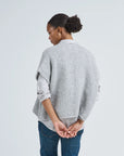 A woman with her hair in a bun stands with her back to the camera, wearing LaTierra’s Flor Vest—a light grey knit made from baby alpaca and merino wool—over a shirt and blue jeans, hands clasped behind her against a plain background.