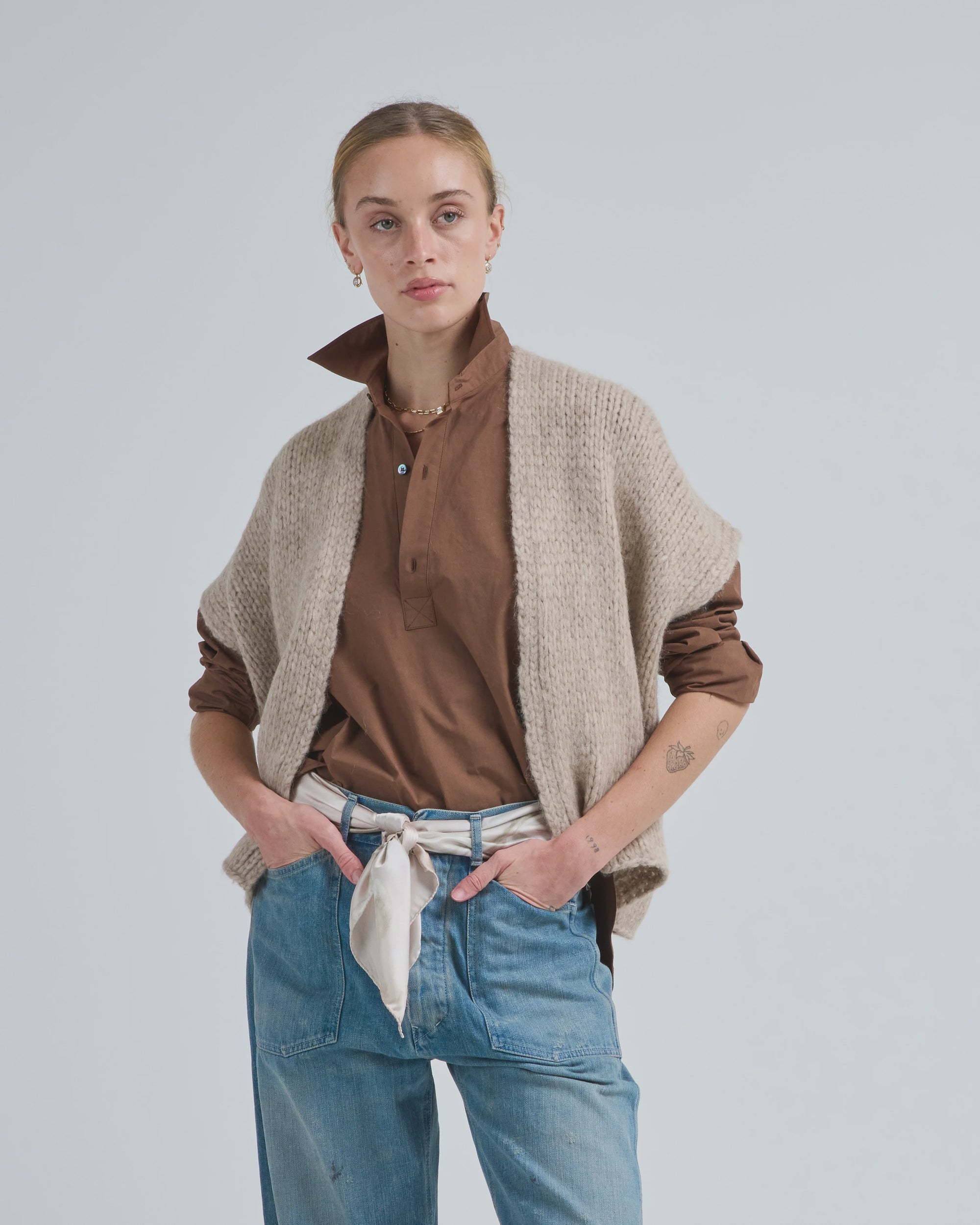 A person with tied-back hair wears the LaTierra Flor Cardigan Vest over a brown shirt and blue jeans with a white scarf belt, standing hands in pockets against a plain light background.