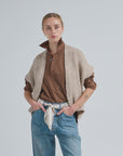 A person with tied-back hair wears the LaTierra Flor Cardigan Vest over a brown shirt and blue jeans with a white scarf belt, standing hands in pockets against a plain light background.