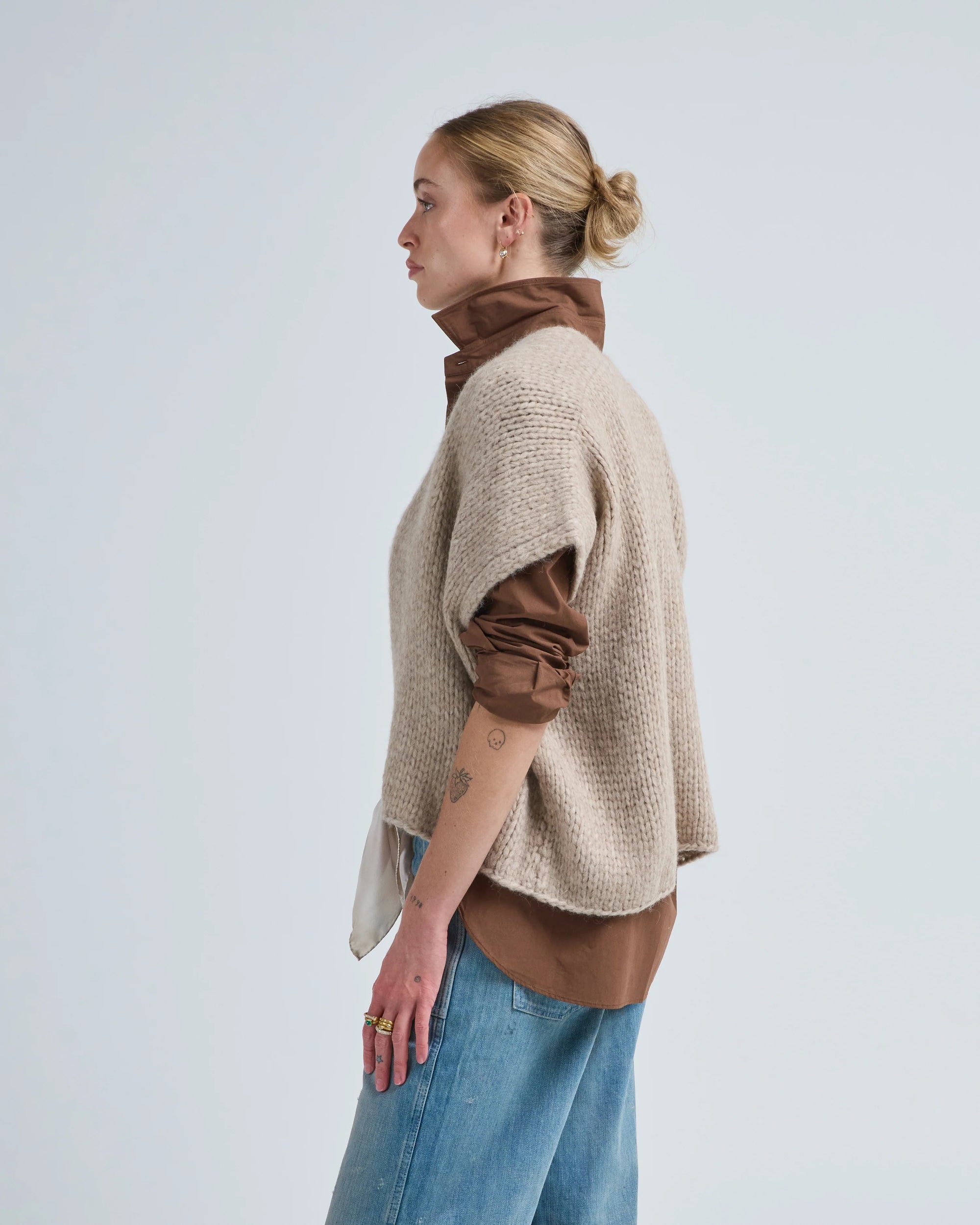 A woman in profile stands against a plain background wearing the LaTierra Flor Cardigan Vest in beige over a brown shirt with rolled-up sleeves and light blue jeans. Her hair is tied back, showing forearm tattoos.
