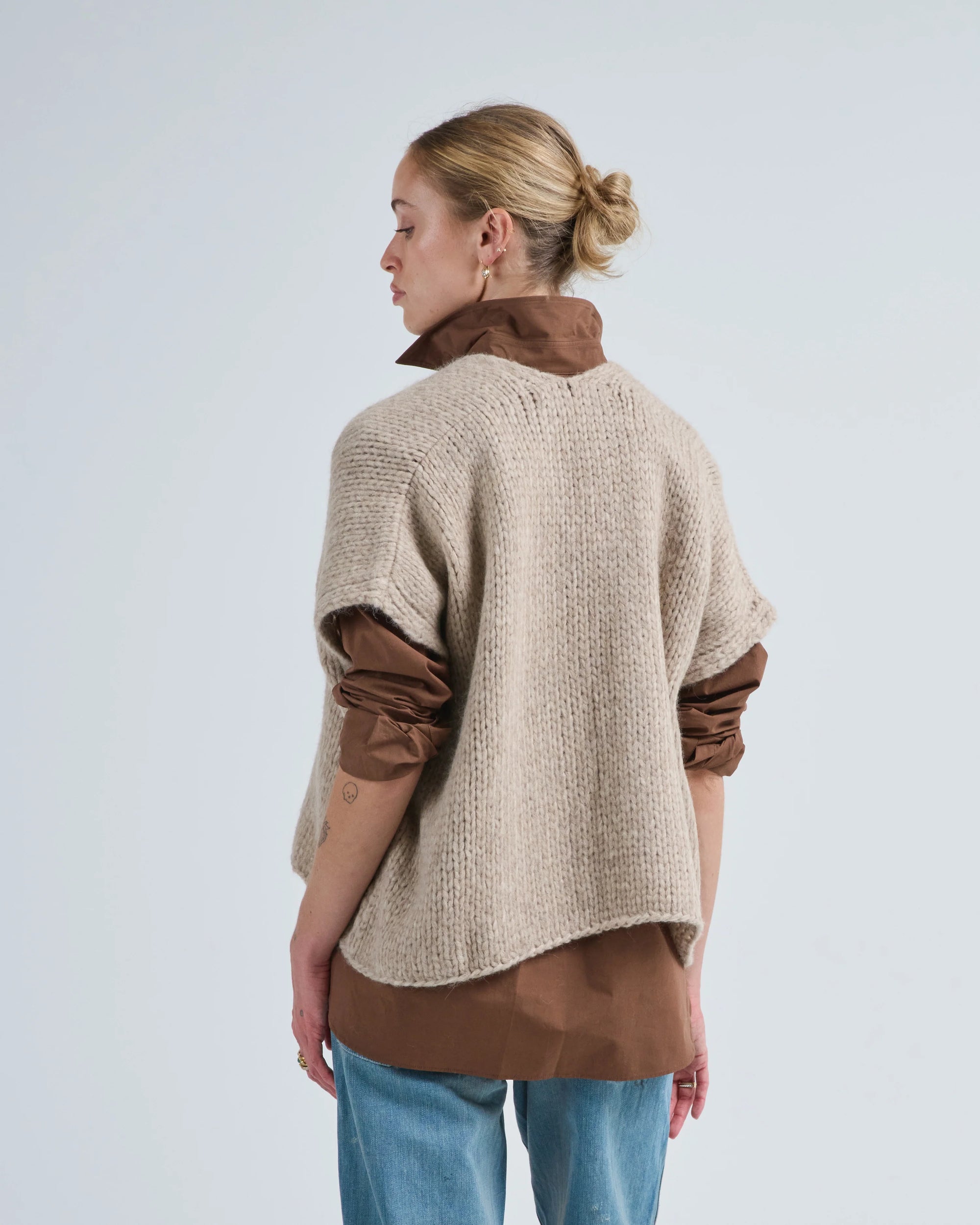 A woman with blonde hair in a bun stands sideways against a plain light background, wearing LaTierra’s Flor Cardigan Vest, a beige merino wool knit, layered over a brown rolled-sleeve shirt and light blue jeans.