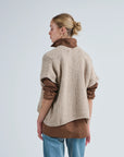 A woman with blonde hair in a bun stands sideways against a plain light background, wearing LaTierra’s Flor Cardigan Vest, a beige merino wool knit, layered over a brown rolled-sleeve shirt and light blue jeans.