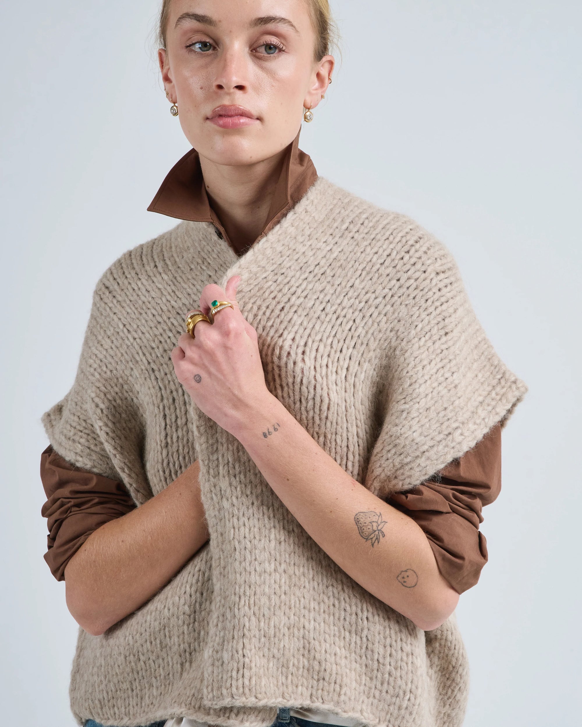 A person with short blond hair, hoop earrings, and forearm tattoos stands against a plain background wearing the LaTierra Flor Cardigan Vest—a chunky beige knit made from soft merino wool—holding it closed with one hand.