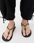 A person in black cuffed pants wears Flowze FLO Sandals—green, rope-style with black soles—while standing on a light-colored surface.
