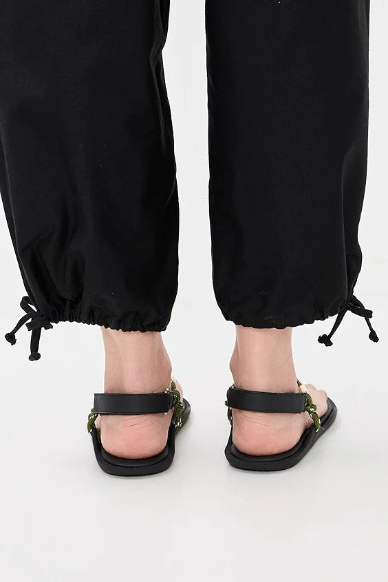 A person wears black drawstring pants and Flowze FLO Sandals—black, lightweight with ankle straps—standing on a white surface. The photo, taken from behind, focuses on the lower legs and feet for an adjustable fit.