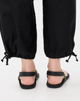 A person wears black drawstring pants and Flowze FLO Sandals—black, lightweight with ankle straps—standing on a white surface. The photo, taken from behind, focuses on the lower legs and feet for an adjustable fit.