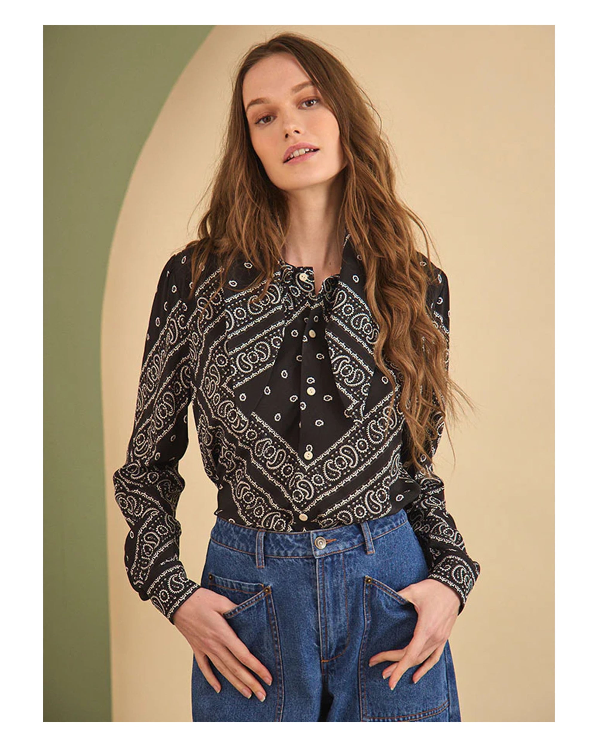 A woman with long wavy brown hair models the Hunter Bell NYC Frances Blouse, a black bandana-print top with a neck bow, paired with high-waisted blue jeans, hands in her pockets against a neutral background.