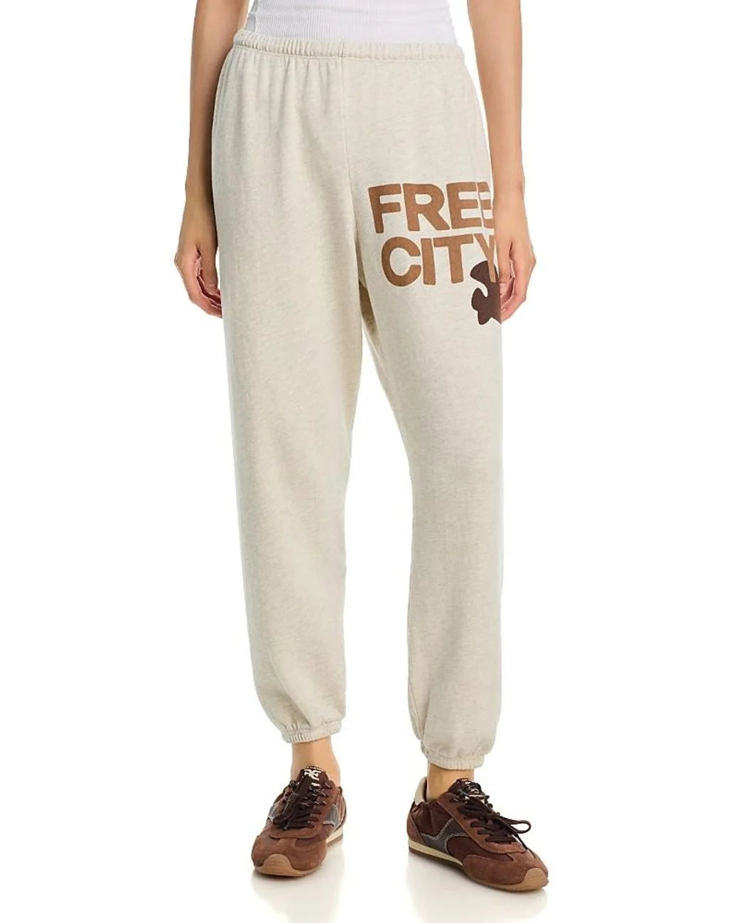 Someone wears the FREECITY Large Sweatpant by Free City (sparrow, LLC) in light beige French terry with "FREE CITY" in bold brown text on the upper left thigh, styled with brown sneakers and a white top.