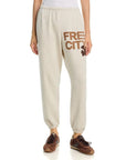 Someone wears the FREECITY Large Sweatpant by Free City (sparrow, LLC) in light beige French terry with "FREE CITY" in bold brown text on the upper left thigh, styled with brown sneakers and a white top.