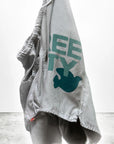 The Free City Matte/Satin BackPocket Short in light gray features frayed edges, teal FREECITY print, and a blue dove graphic, shown hanging against a plain background.