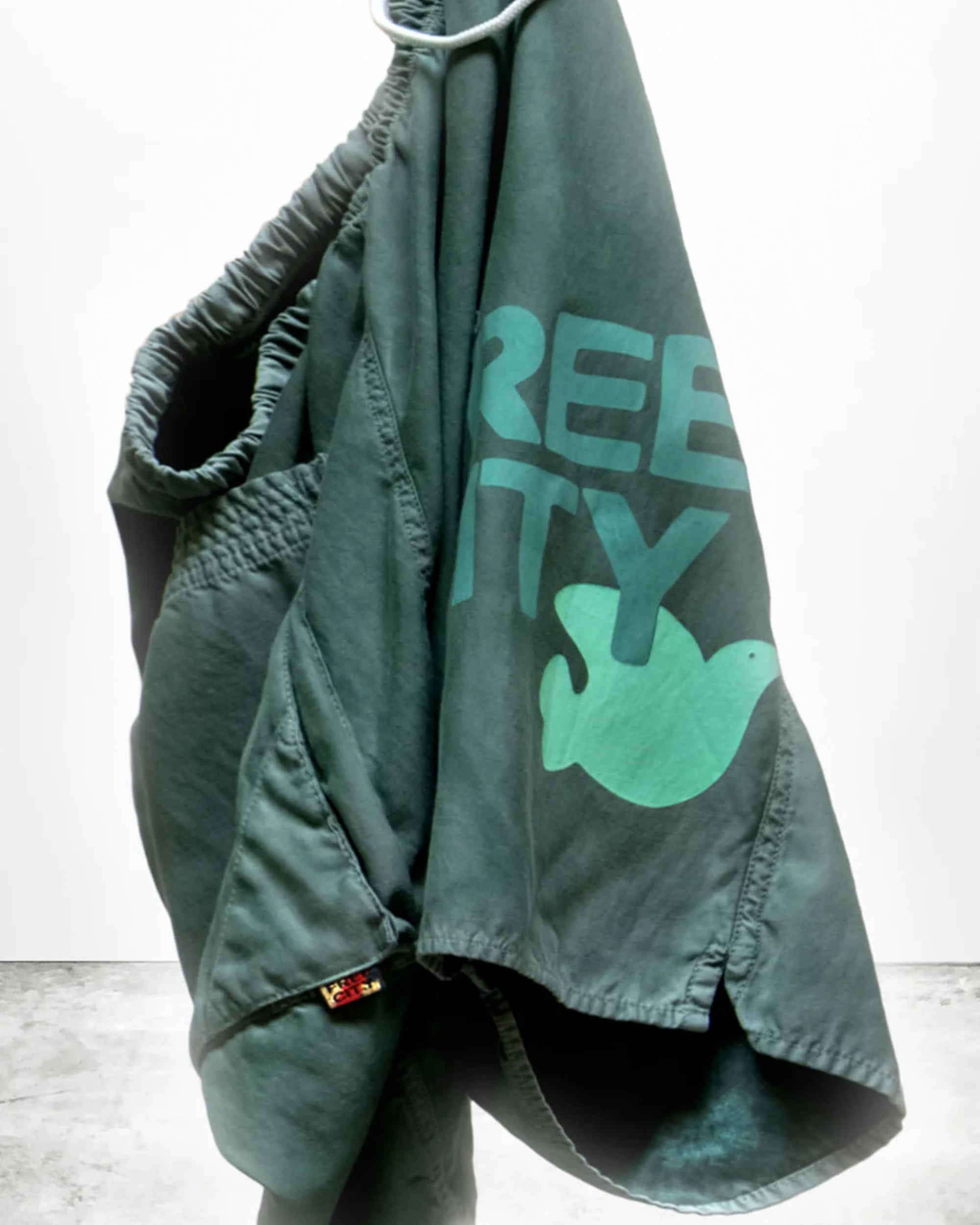 A pair of green Free City Matte/Satin BackPocket Shorts with an elastic waistband hangs on a hook. Handmade in Los Angeles, these shorts feature a dove graphic and partial "REE TY" text in light green.