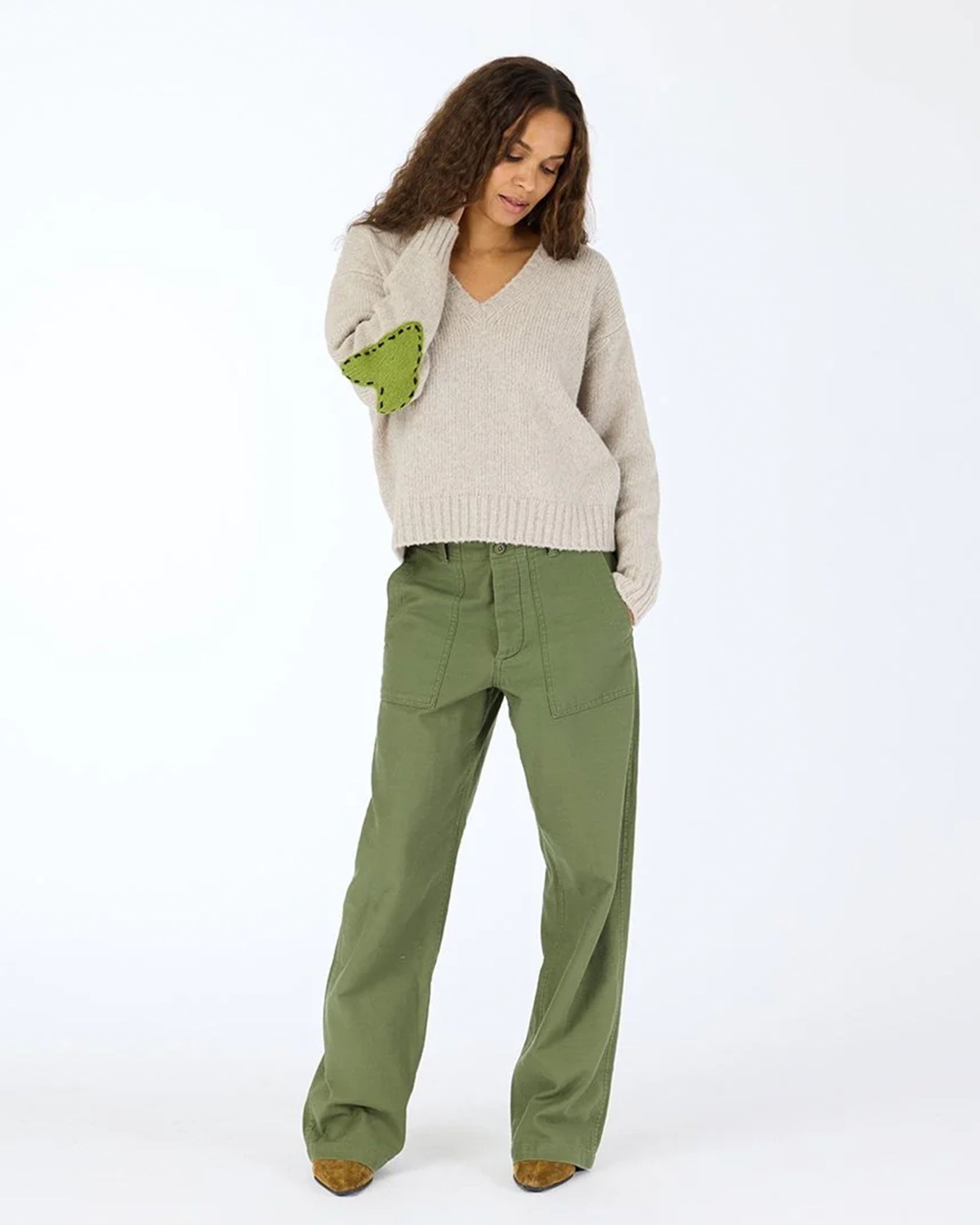A woman with curly hair wears the Kerri Rosenthal Frenchy Sweater with heart patch elbows and relaxed-fit olive green cargo pants, standing against a plain white background.