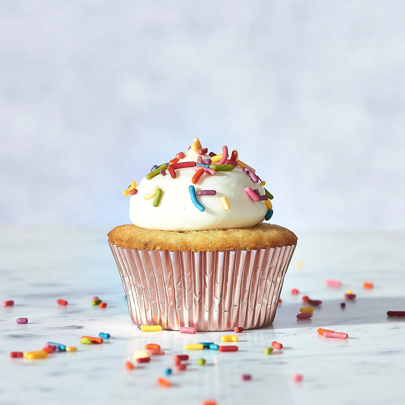A Wild Rye Frosting plant-based vanilla cupcake with white vanilla frosting and colorful sprinkles sits in a shiny pink wrapper on a marble surface, with extra sprinkles scattered around.
