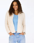 A woman with long curly hair smiles, hands in her pockets, wearing the Kerri Rosenthal Fringe Beaulero Cardigan in cream over a light blue top and light-wash jeans, standing against a plain white background.