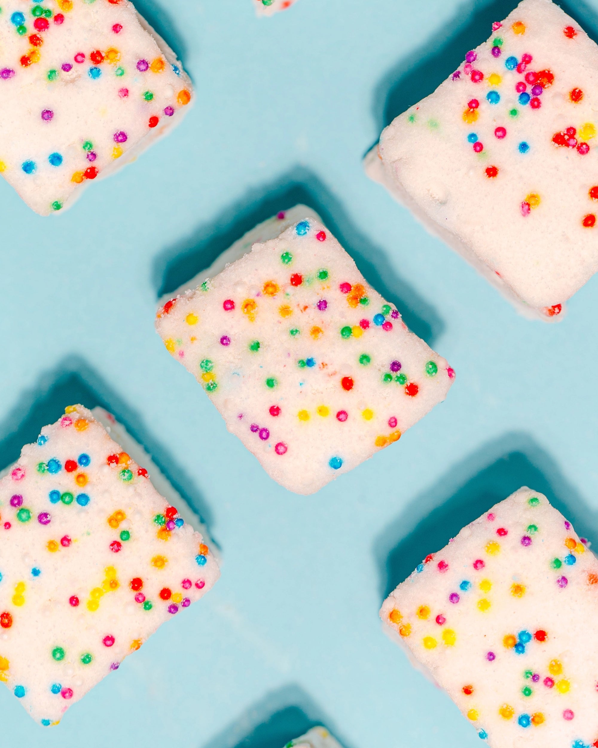 Faire Funfetti Marshmallows—colorful, allergy-friendly squares with rainbow sprinkles—are evenly spaced on a light blue background in a playful diagonal pattern.