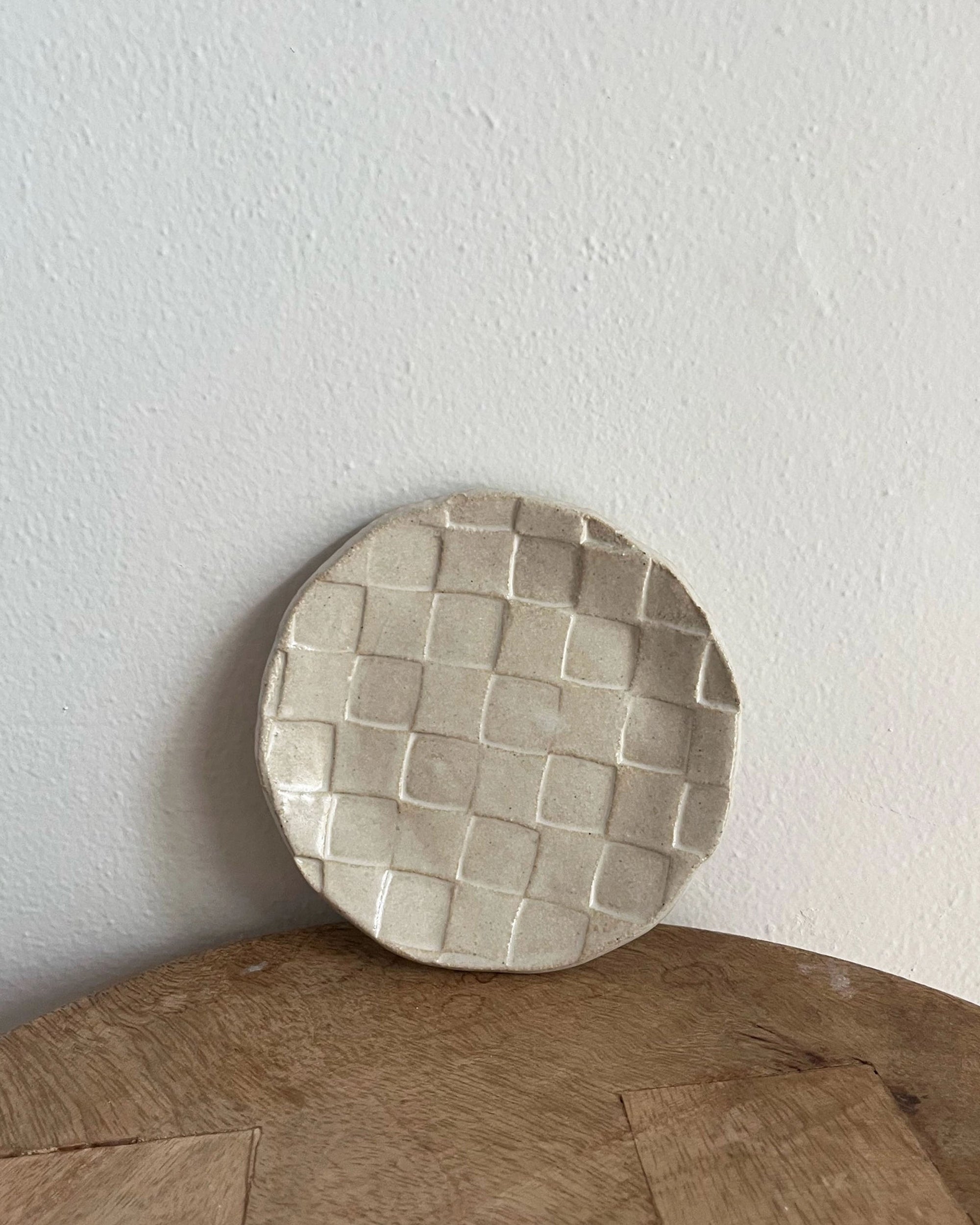 The Faire Ghost Checker Trinket Dish is a round glazed stoneware plate featuring a light beige checker pattern, displayed upright against a white wall on a wooden surface.