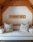 A "Giddy Up" Felt Banner by Faire hangs above a cozy couch with two patterned pillows in a wooden nook, featuring horse, cowboy, and cactus accents—an eye-catching cowboy gift idea.