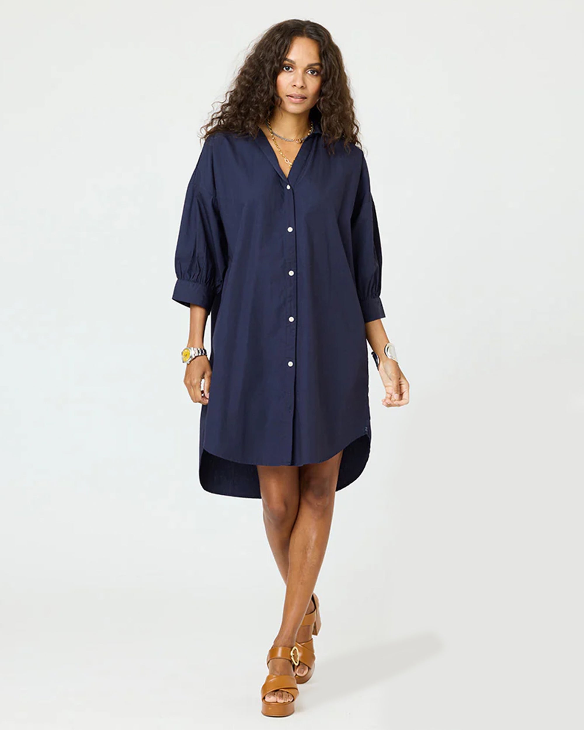 A woman in the Kerri Rosenthal Gigi Denim Dress—loose-fitting, knee-length, navy blue, 100% cotton with three-quarter sleeves—pairs it with brown sandals and stands against a plain light background.