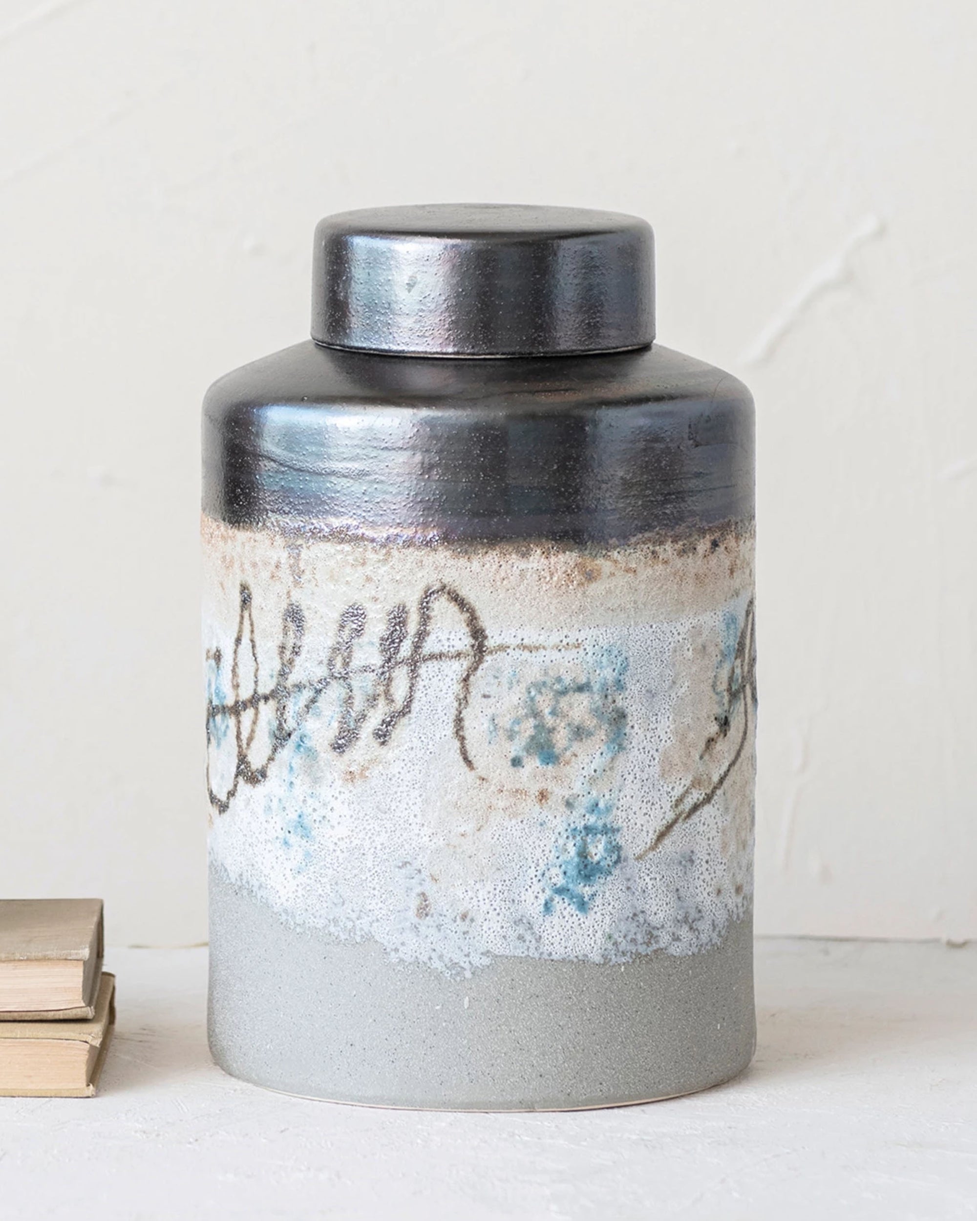 The Creative Co-op Ginger Jar is hand-painted stoneware with a round lid, showcasing blue, gray, and beige tones. Abstract wavy lines and subtle floral motifs accent its textured surface. It rests on a light surface beside stacked books.