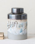 The Creative Co-op Ginger Jar is hand-painted stoneware with a round lid, showcasing blue, gray, and beige tones. Abstract wavy lines and subtle floral motifs accent its textured surface. It rests on a light surface beside stacked books.