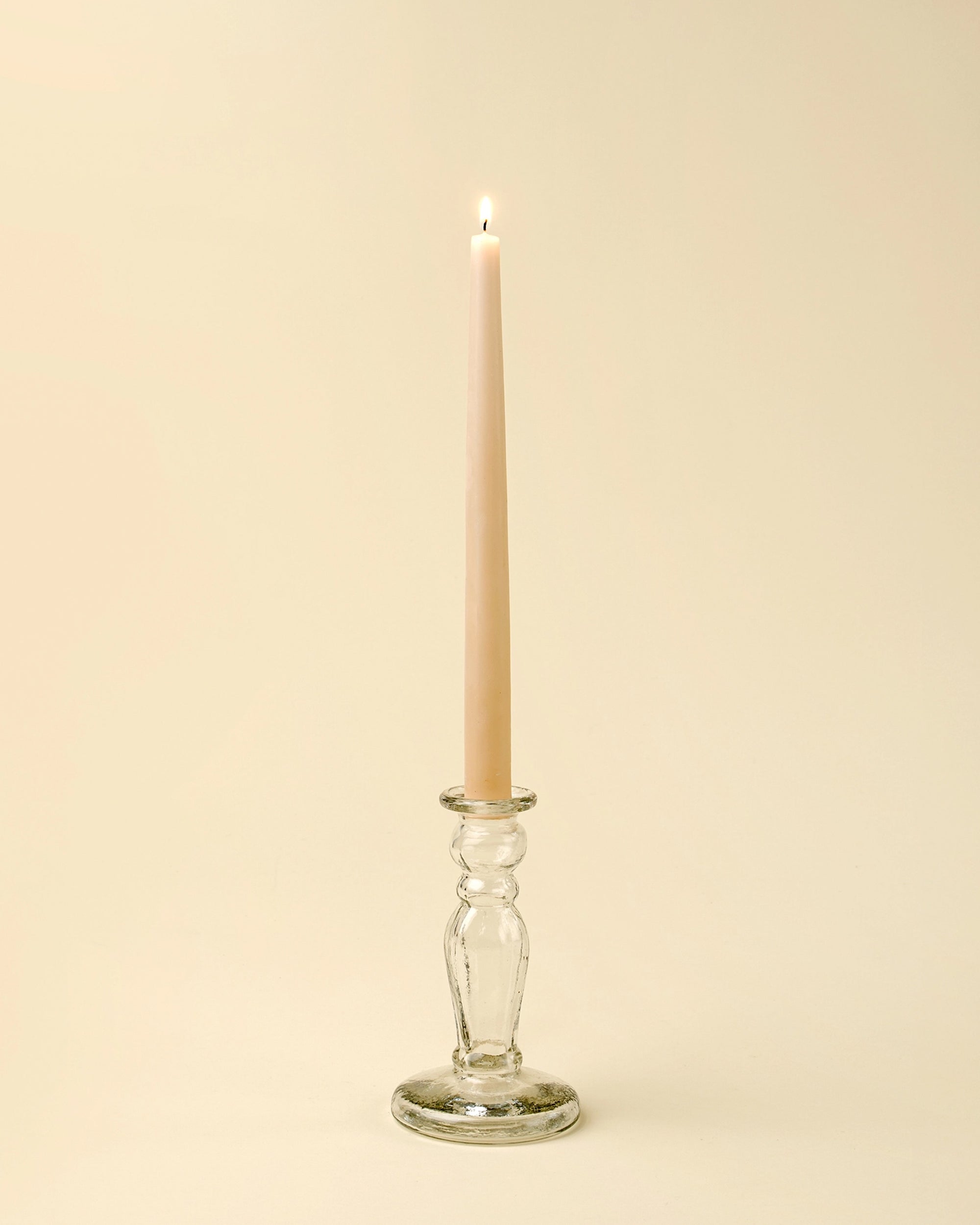 A lit beige taper candle stands upright in the Indaba Vero Glass Candlestick S, crafted from handblown recycled glass, set against a plain, light cream background.